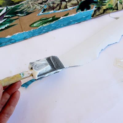 A foam brush applying modge podge to the backside of an illustration from The Little Island.