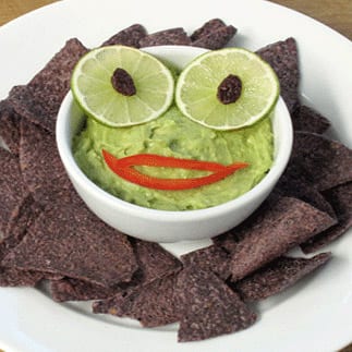 Bowl of guacamole with lime slices and raisins for eyes and red pepper slices for a mouth surrounded by blue corn tortilla chips.